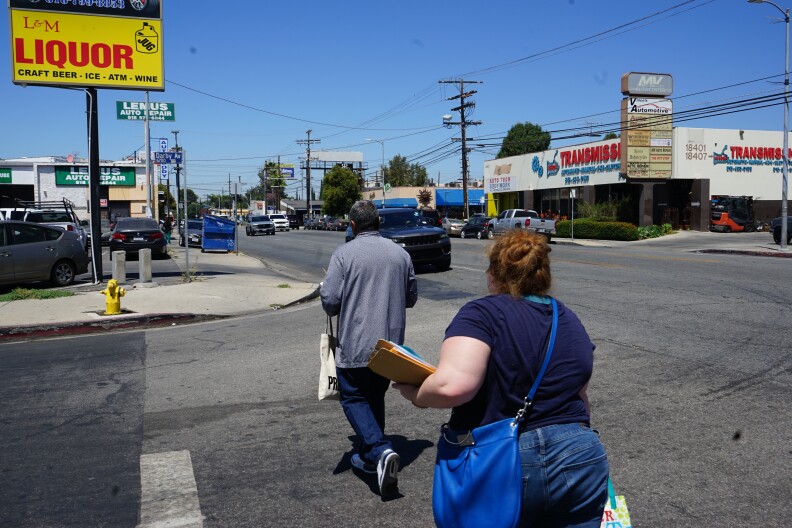 Two people cross a street on a bright, sunny day. The sky is blue. They walk towards an auto repair shop. A transmission shop is across the street. Cars are driving by on the street. The person in front has a buzz cut, gray jacket and jeans. Behind him, a woman with light skin and red hair in a bun follows, carrying clipboards.