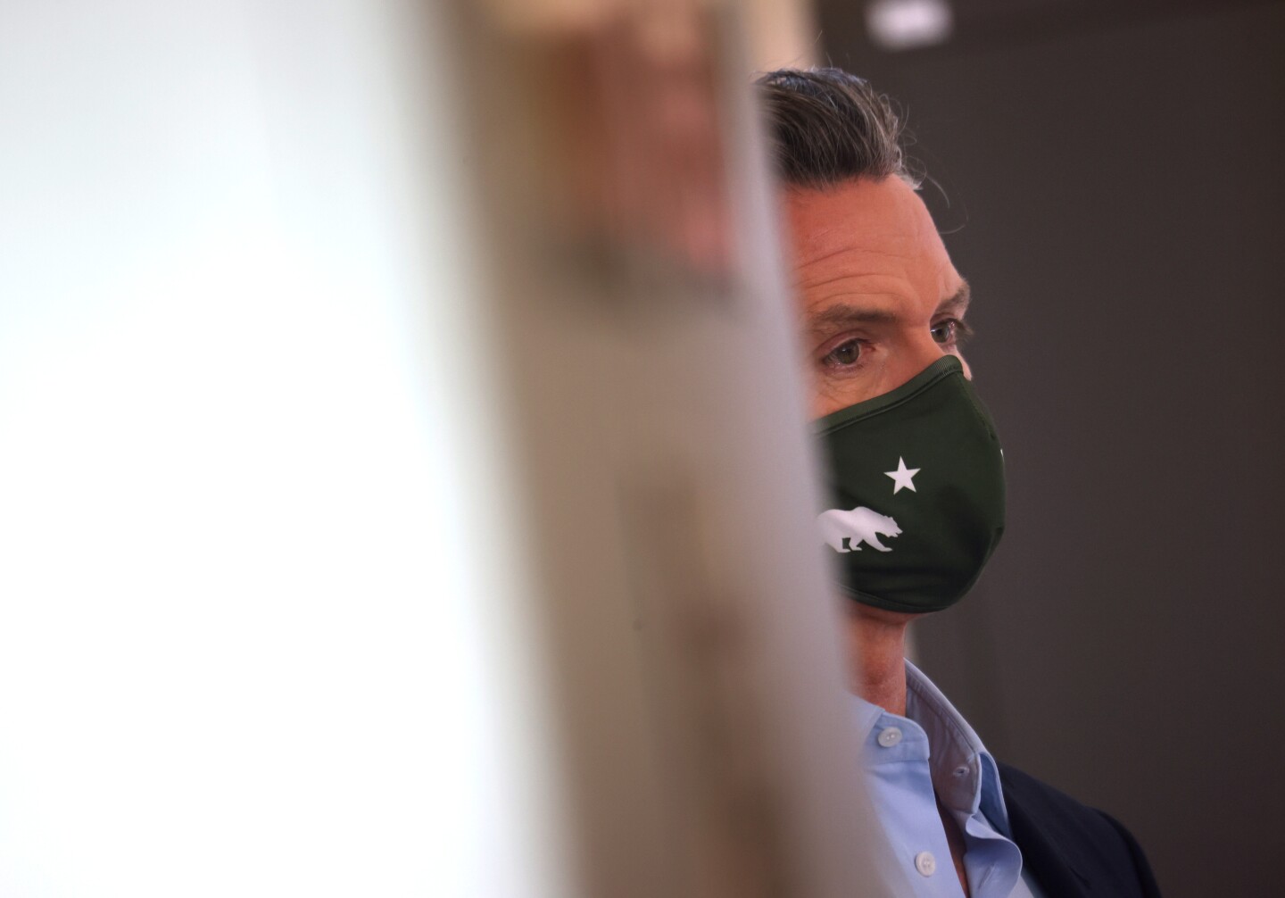Governor Gavin Newsom, in profile, waits to speak at a news conference while wearing a black mask with the state's bear in white.