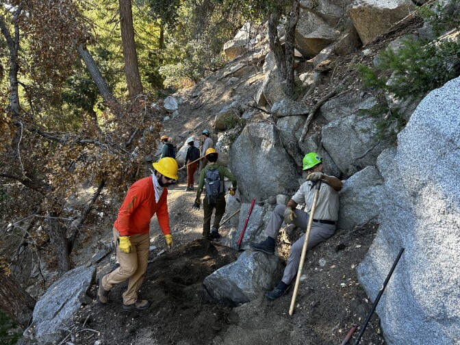 Multiple younger and middle-aged men on a dirt trail with large boulders wearing hardhats and holding McLeod tools do work on a trail. 