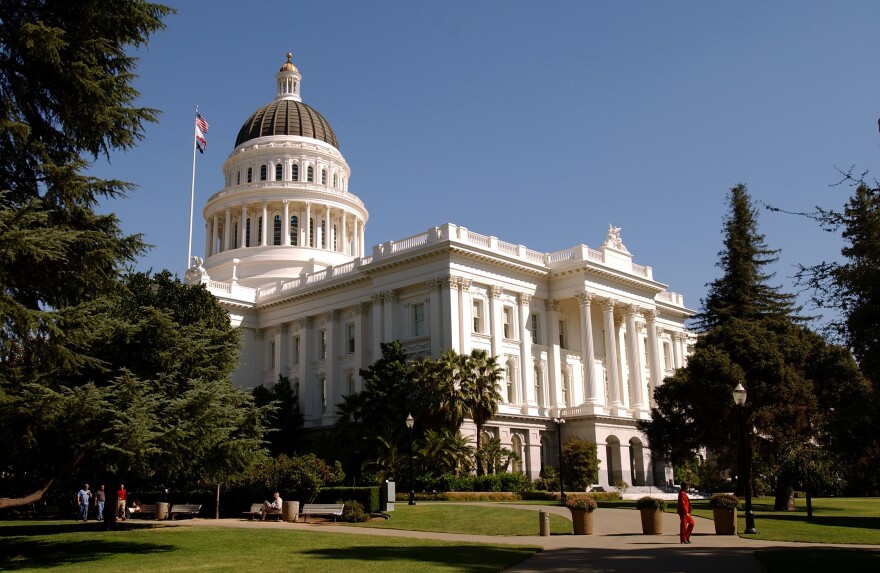 An image of a large building with a dome on top of it, the California capitol building, with evergreen trees nearby seen in the foreground.