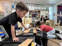  A student with light skin tone and black track pants and t-shirt picks up a wooden track. In the background, students with a variety of skin tones look at other parts of the track. 