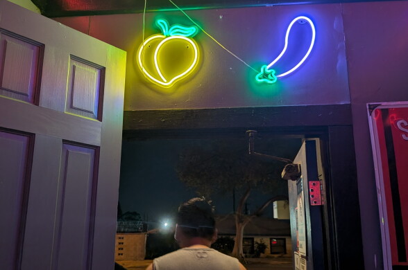 A person walks out of a door into the streets at night. Above the door are two neon signs of a beach and a cucumber.