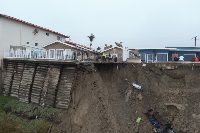 The collapse this week cut away part of a patio where several people can be seen standing. What looks like patio furniture and other construction materials have slid down the hillside along with the dirt. Another portion of the cliff is reinforced with wooden scaffolding. 