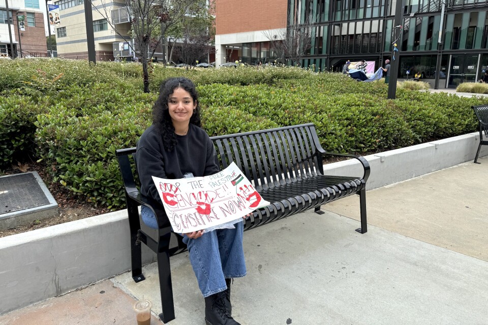 A young woman with light brown skin sits on an outdoor bench holding a sign that says "ceasefire now."