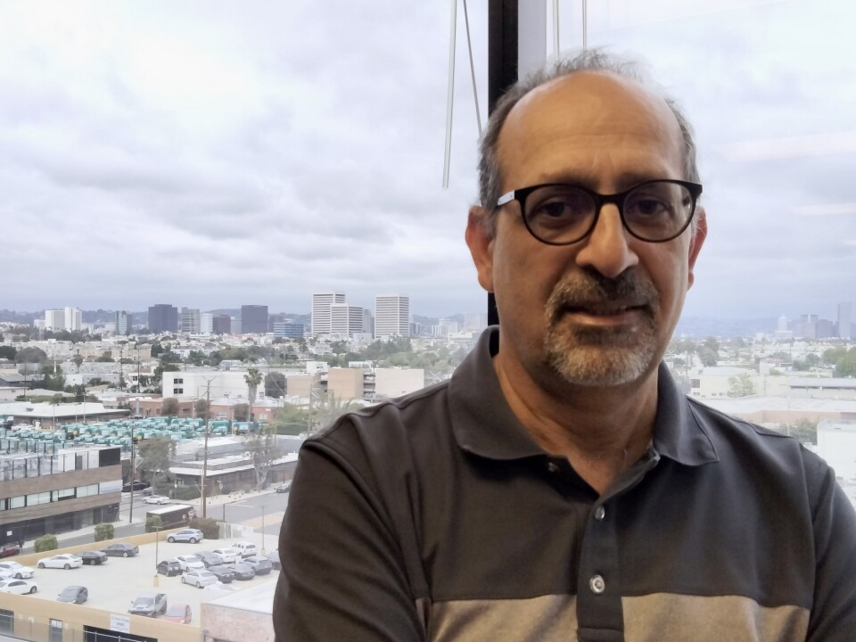 Man with medium-brown skin, glasses and two-tone polo shirt. In the background are medium sized buildings