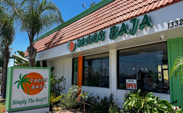 The building exterior features a slanted roof with green lettering signage that reads "Tacos Baja." Below the sign, there are two windows—one with an open sign. The building is surrounded by lush green foliage. In the foreground, there is a standalone sign that reads "Tacos Baja, Simply The Best."