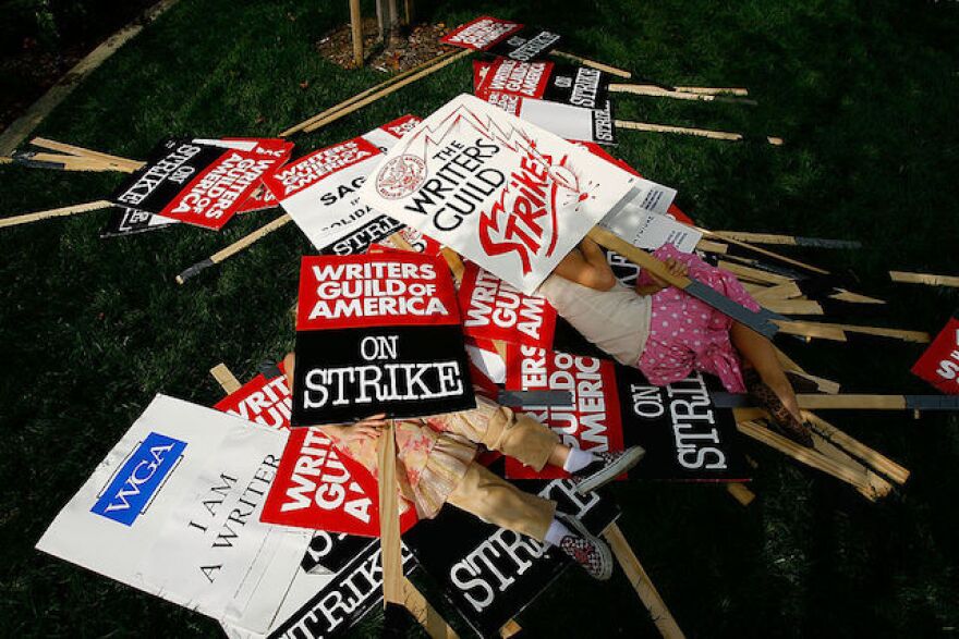 A variety of signs with wooden handles created by the Writers Guild of America in a pile. Several signs say, "Writers Guild of America: On Strike"