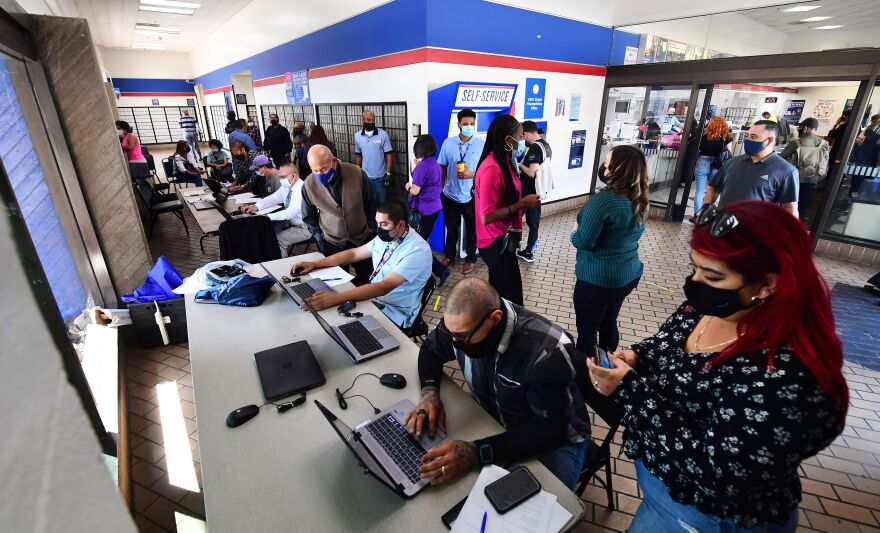 People wearing masks sit at tables using computers while other people stand around them inside a post office.