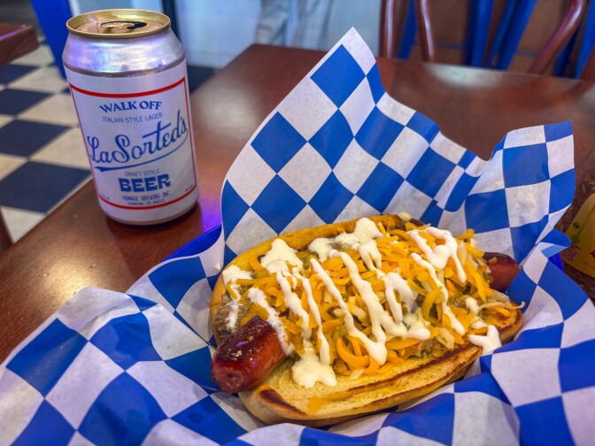 A hot dog in a grilled yellow bun, topped with green sauce, orange cheese, and white cream, rests on a blue and white checkered wrapper. Next to it is a can of "Walk Off Italian Style Lager" beer on a brown wooden tabletop. 