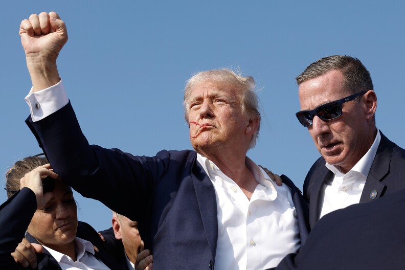 A man with light-tone skin and gray hair raises his right fist as blood trickles down his face. Two secret service agents have arms around him