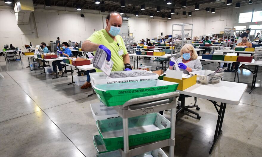 A wide shot of an election worker, who's a person with a medium-light skin tone, wears a mask and gloves as they pull out a batch of mail-in ballots from a united states postal service cart. In the background are rows of white tables with workers at them with trays for ballot processing.