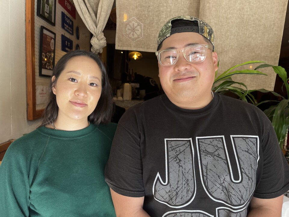 An Asian American woman in a green shirt and Asian American man in a black t-shirt pose for a picture inside a Japanese restaurant.