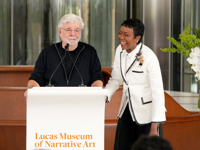 A person speaks at a podium reading 'Lucas Museum of Narrative Art' as another person laughs beside them.