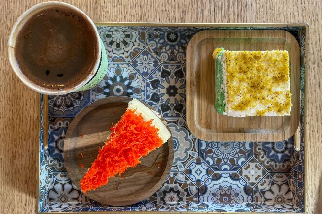 A tray with a blue-tiled design sits on a light wooden tabletop. The tray includes a large white paper cup filled with dark brown coffee with a small amount of brown foam gathered on the top. In the bottom corner of the tray is a small circular wooden plate with a triangular slice of white cheesecake topped with an orange-spun pastry topping. In the upper right-hand corner sits a square cake with a light green crumb at the bottom containing white frosting and bits of a yellow-light-green topping. 