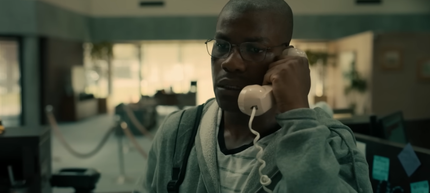 John Boyega is seen from the shoulders up. He looks disheveled with a white t-shirt with thin grey stripes on him that has a neck that is stretched out. Over that, he has a plain gray hoodie that has shifted on his shoulders to be uneven. He has a shaved head with the inklings of new hari growth, and wears a pair of squared oval glasses. He has a landline phone picked up and is holding it to his face. in the background: an empty bank.