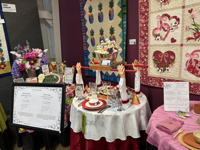 A display of round tables with quilts in the background. The one on the far left features a colorful floral centerpiece and the one in the center features four mannequin arms holding a giant faux ice cream sundae.
