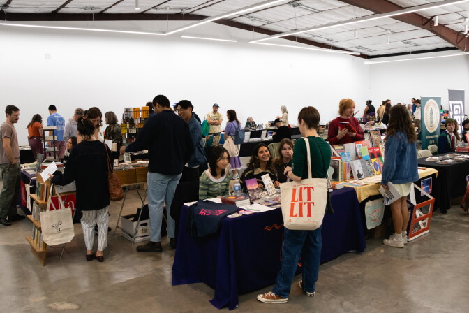 A crowd of people stand in a room with different tables. Books are displayed on the tables. The ground is concrete and grey. A person in the foreground carries a tote bag that says "LITLIT"