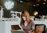 A fair skinned woman with red hair and tattoos sits in a coffee shop with laptop.