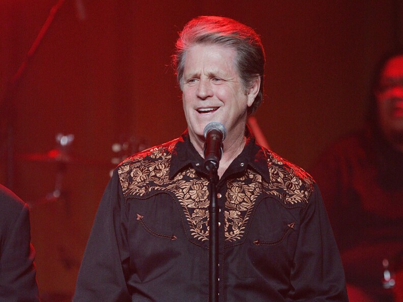 An older white man in a dark button up shirt sings into a microphone.