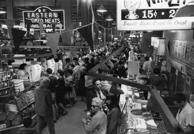 A black-and-white photo offers a long-shot view of an open air market: A sign overhead declares "Eastern Smoked Meats Hams Bacon" and another snack bar sign announces deals for 15 cents. Many women are dressed for shopping, in knee-length skirts, while the skinny ties and jackets and cat eye sunglasses offer a sense of the time period. 