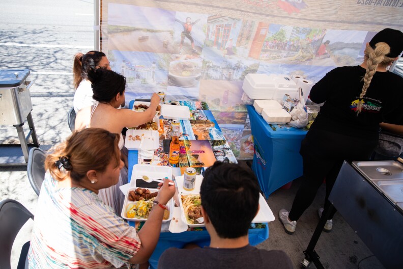 Various people sit under a pop-up tent at a long table with a blue table cloth full of styrofoam boxes with food.