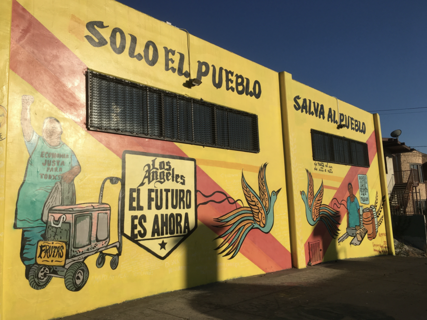 A brightly colored mural with a yellow backdrop depicts a street vendor with a pushcart and a raised fist.