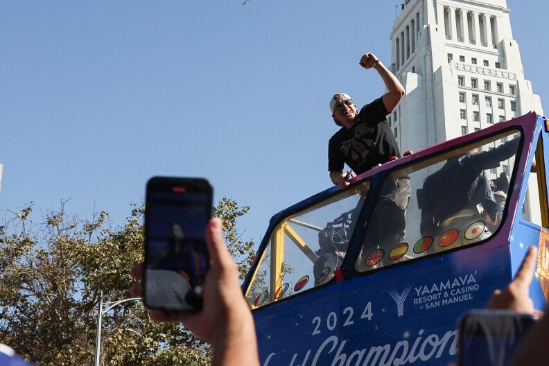 A man wearing a black shirt, white baseball cap, and sunglasses leans against a double decker bus and raises a fist in celebration