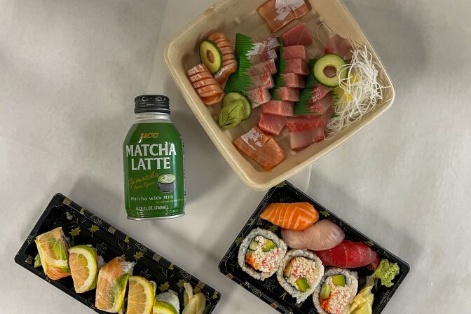 Three containers of different types of sushi were placed on a white background. On the bottom left is a roll containing thinly sliced lemon topped with pink salmon and avocado. The container next to it contains three large pieces of cut California roll sushi along with three pieces of sushi containing pink salmon,  white escolar, and red tuna. Above both is a large square container featuring various cuts of fish arranged next to each other. Between the containers is a short aluminum bottle with a green label and black top with the words 'Matcha Latte' printed.
