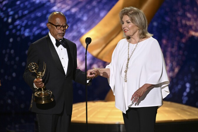 Two people appear on a stage. On the left is a man wearing a tuxedo, holding a statuette in his right hand. To his left a woman wearing a white blouse and long gold chain stands next to him, they are holding hands. In front of them is a long, slender microphone.