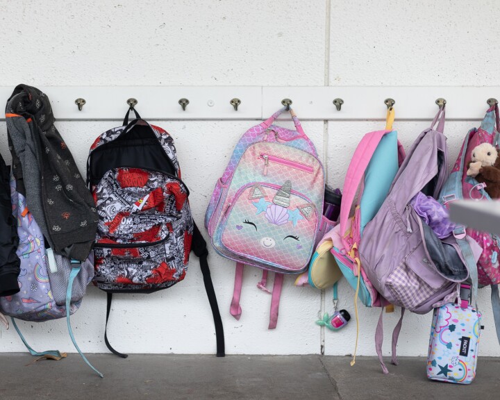 A white wall with metal hangers lined with children's school backpacks of various colors.