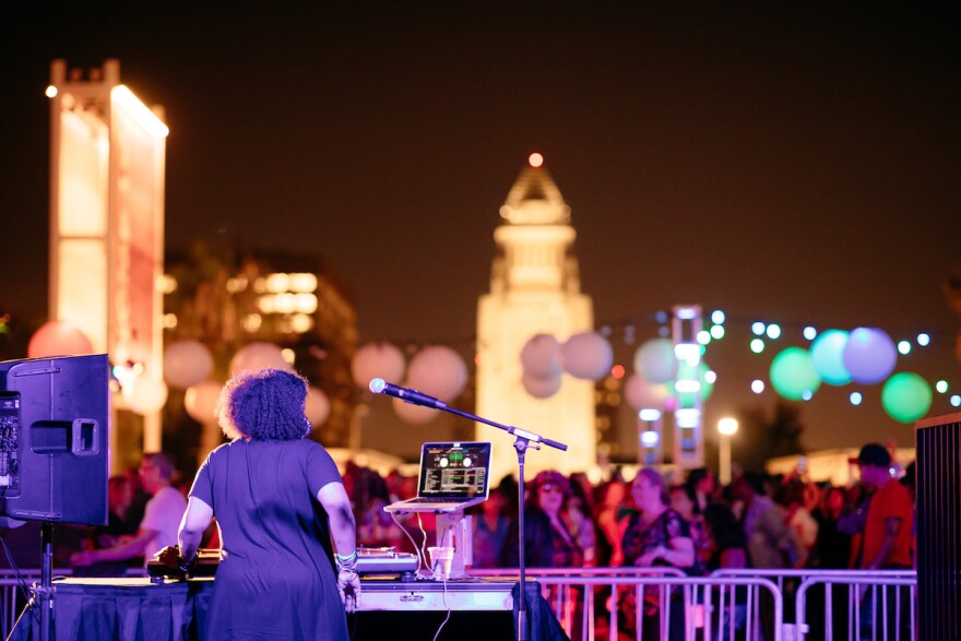 A night time image of a woman with her back to the camera DJing on a stage as a crowd watches. LA City Hall is lit up in the background