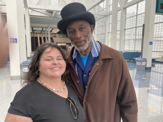 A stout white woman with a green top stands pressed against a Black man who is several inches taller than her. He is wearing a brown jacket and a black top hat. He has a white beard.