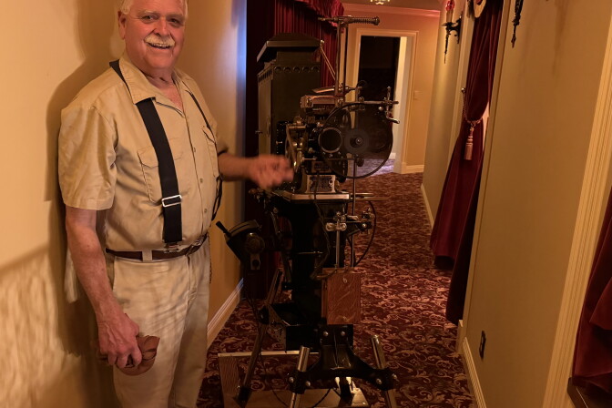 An older man with a gray mustache wearing a tan shirt, white pants and suspenders is handcranking an antique movie projector