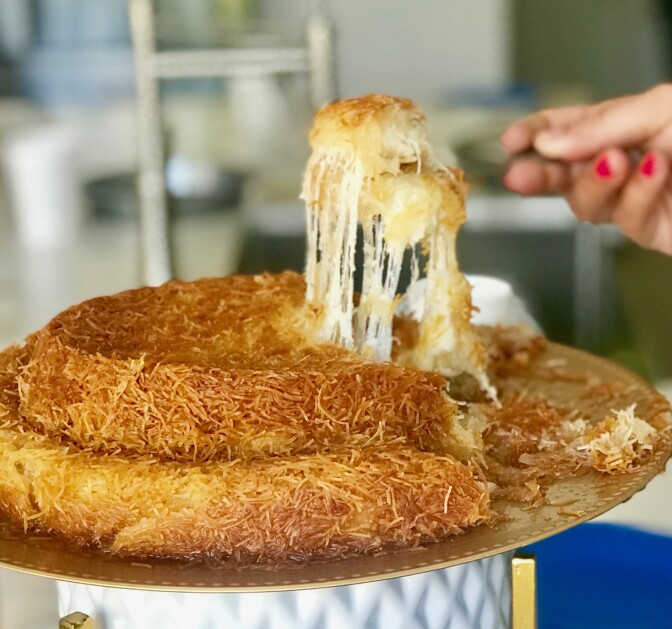 A two layer shredded phyllo dough and cheese dessert on a gold tray. A hand with red nailpolish painted on the nails lifts up a slice. 