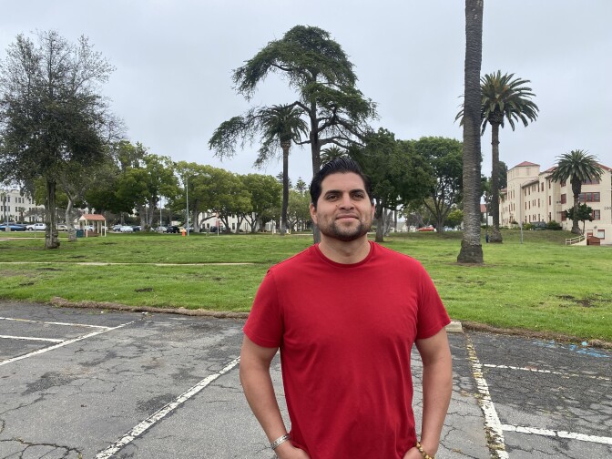  A dark haired man with a short beard in a red T-shirt smiles while standing on an asphalt lot in front of grass, a mix of trees and an older off-white stucco building on the West LA campus 