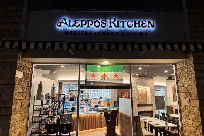 A black, green and white flag hangs outside a restaurant. The words Aleppo's Kitchen are backlit on the restaurant sign.