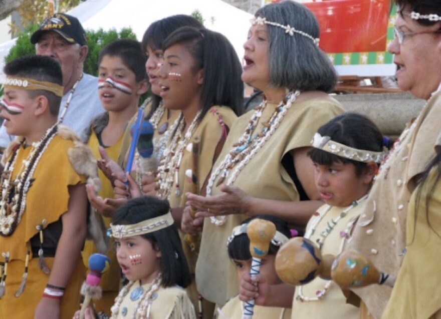 A line of 10 people, one man, two women, and seven children stand mostly shoulder to shoulder, dressed in traditional tan and yellow Tongva clothes, including shell necklaces and headbands, with several holding Tongva percussion instruments, with their mouths open as if they are signing or chanting. Five of the kids wear face paint; two of the boys' face paint consists of a horizontal stack of red, white and blue stripes running from one cheek, over their noses and on to their other cheek.