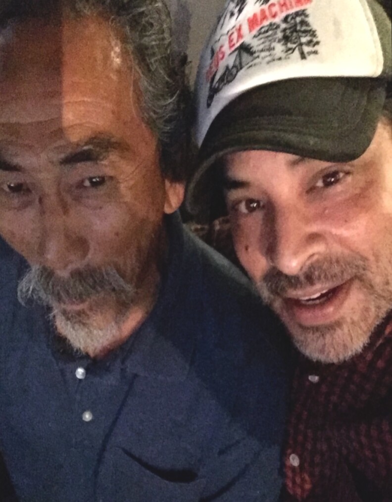 A gray-haired man with a goatee poses with his son who is wearing a baseball cap.