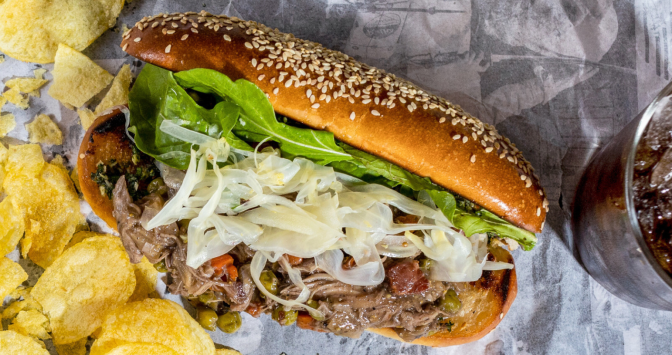 A sandwich has a small bun filled with shredded cooked meat, thin slices of white onion, and leafy green lettuce. On the left side is a portion of yellow potato chips, and on the right is a glass of cola.
