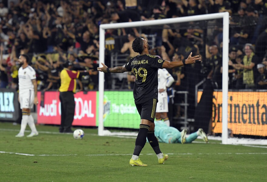 A soccer player with dark-toned skin raises his arms to either side and looks upward in front of goal.