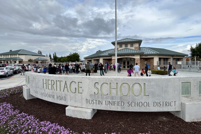 A long, low concrete structure reads Heritage School, and below it, Tustin Unified School District. Behind the structure, a large group of kids and adults stand and walk in front of a building with a green roof that reads "Administration."