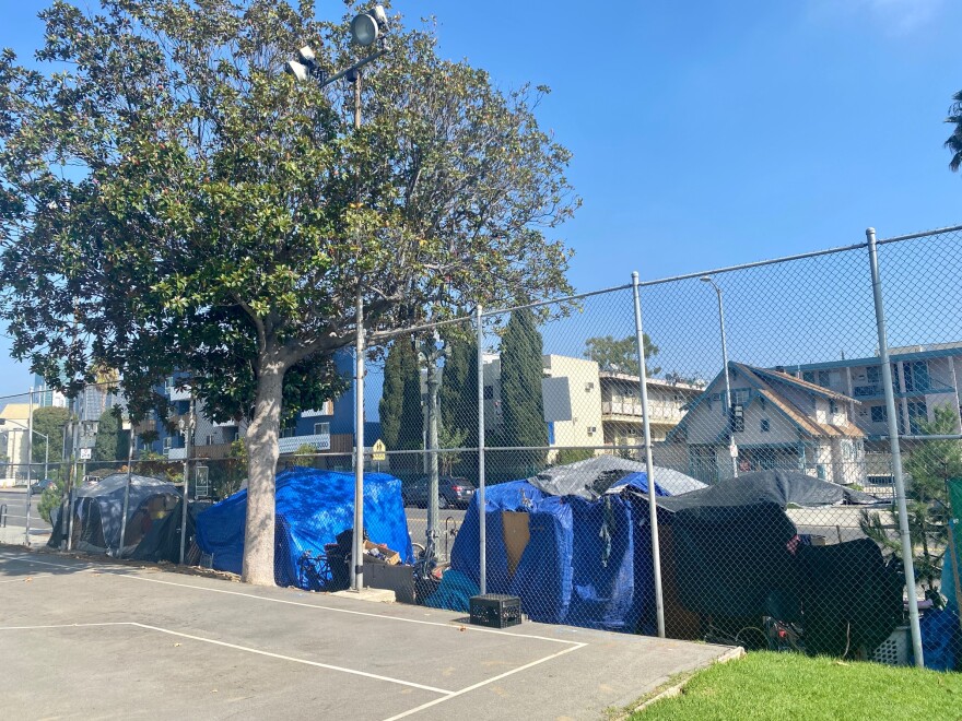 An encampment of tents at the Hollywood Recreation Center lines the street. The photo is taken from inside the park and the tents are shown on the other side of the fence, with apartment buildings across the street.