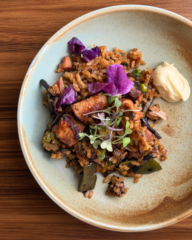 A close-up overhead shot of a speckled ceramic bowl holding a portion of rice studded with charred vegetables and browned meat, garnished with purple flower petals and microgreens. A dollop of pale yellow aioli sits beside the rice on the same plate, against a dark wood table.