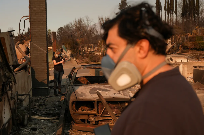 Blurred in the foreground is a profile of a person wearing a mask looking to the left side. Behind them is another person on their phone wearing a mask and standing between a burned car and building. In the background are burned trees and buildings.