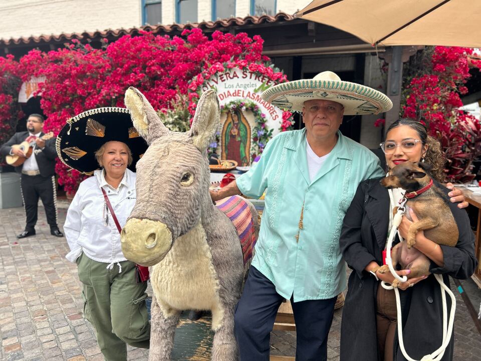 A portrait of three people with medium light skin tones standing near the stuffed donkey. On the sides are two women, who are smiling for the photo. One is holding a dog and the other has a sombrero on. In the middle is Richard, a man with a sombrero on as he puts his hand on the donkey.