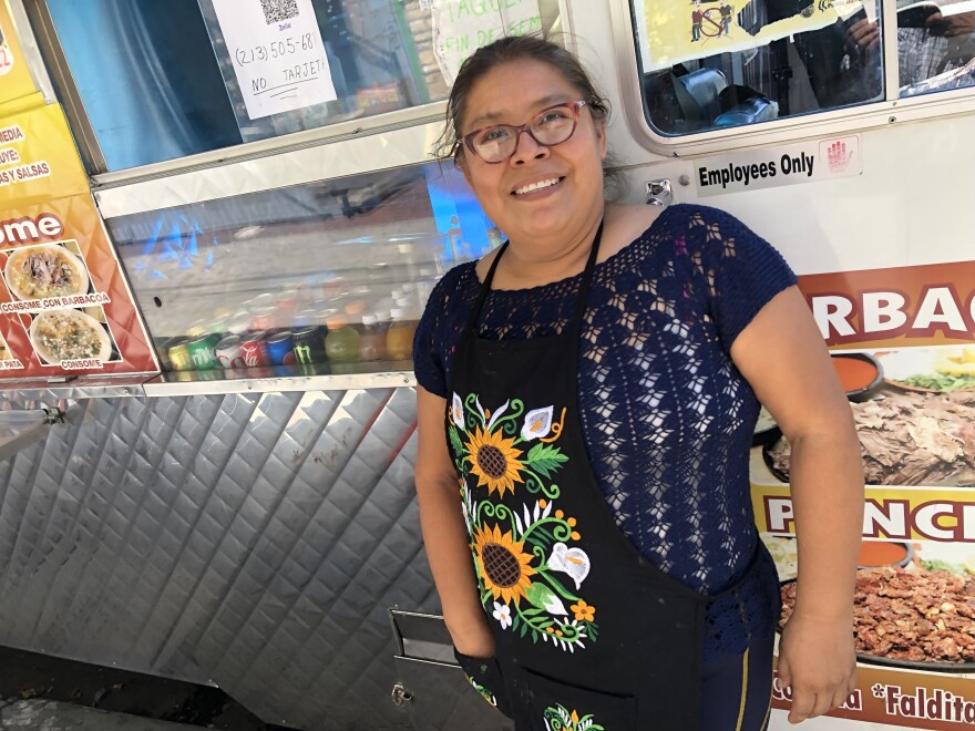 Woman food truck worker is dressed in sunflower apron and stands in front of her food truck that sells Mexican barbacoa and other items.