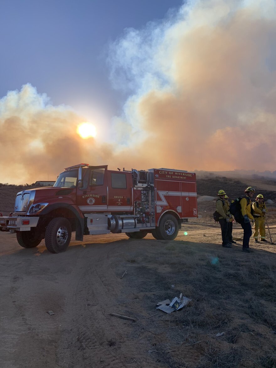 A firefighting truck with personnel standing next to it. Smoke clouds are seen in the background.