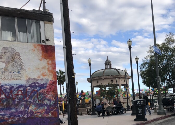 To the left a wall has a mural of a woman with graffiti over some landscape. To the right a park square features an elaborate  gazebo. People are walking through the park.