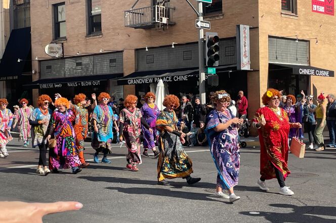 A group of women in orange curly wigs and colorful print dresses, in the style of the character Helen Roper from "Three's Company," walk down the street in a parade.