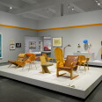 Gallery view of mid-century modern chairs in blond wood and leather displayed on a white platform, with two blue anatomical blueprint-style drawings on the left wall and a colorful modular shelving unit and lamps on a pedestal at right.